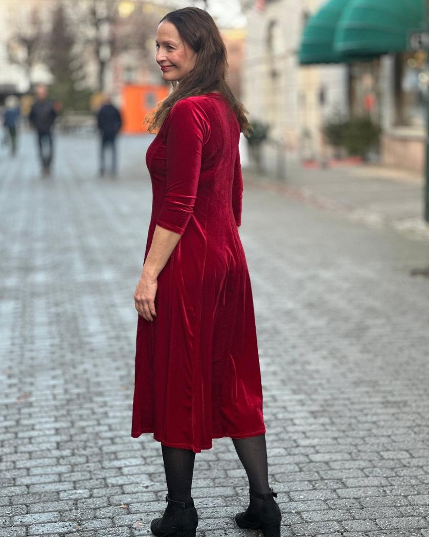 Kvinne i dyp rød velourkjole med V-hals og trekvart ermer, står på brosteinsgate.