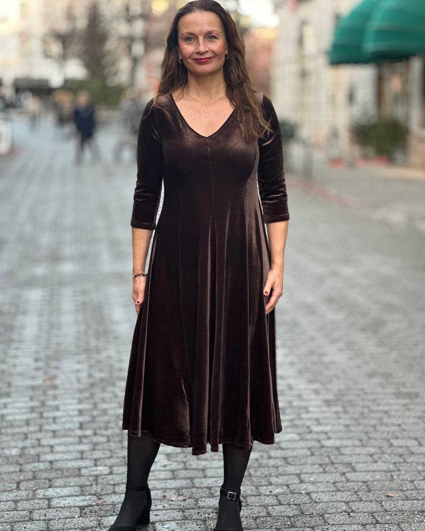 Kvinne i brun velourkjole med V-hals, trekvartlange ermer og A-linjeform, på brosteinsgate.
