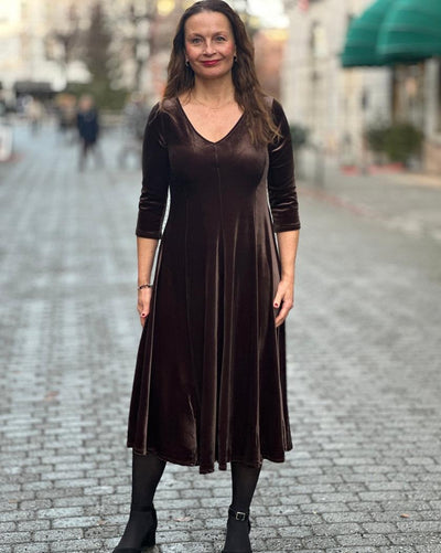 Kvinne i brun velourkjole med V-hals, trekvartlange ermer og A-linjeform, på brosteinsgate.