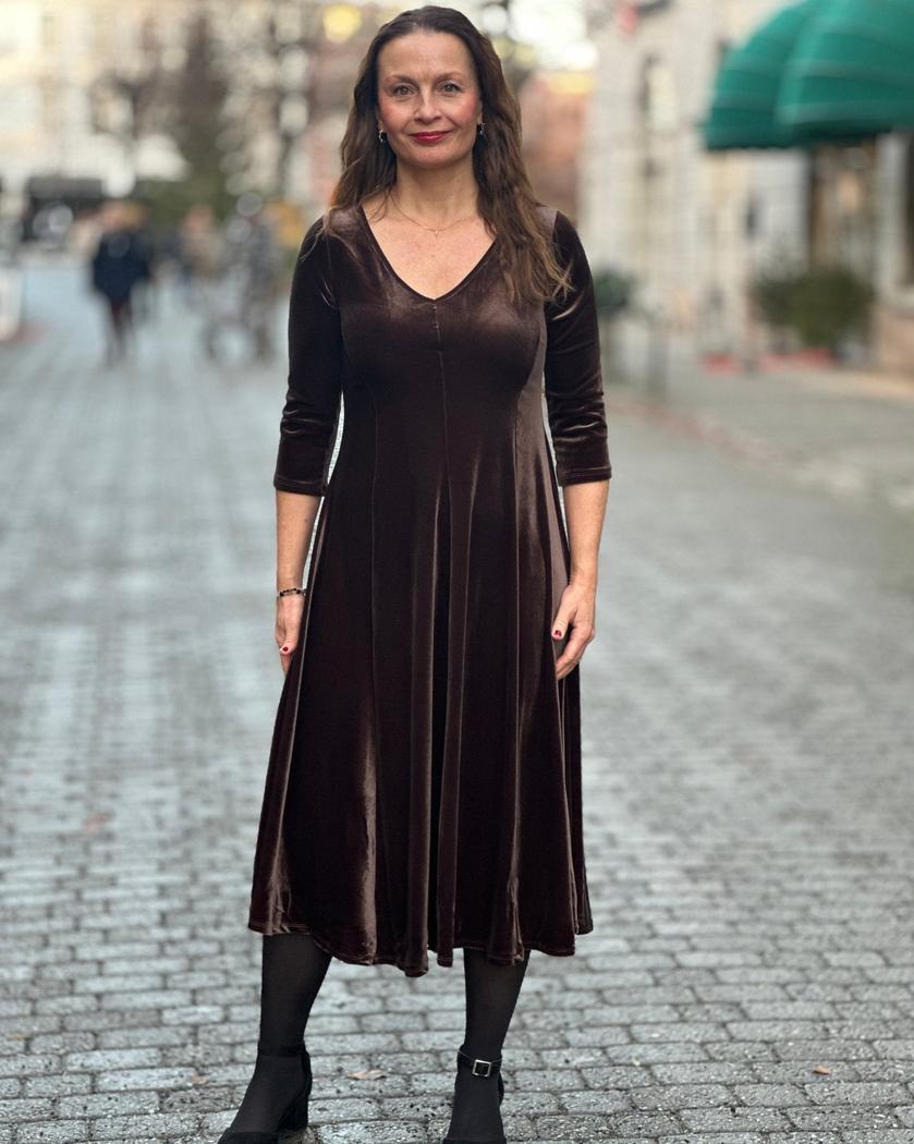 Brun velourkjole med V-hals, trekvart ermer, A-linjeform. Kvinne står på brosteinsgate.