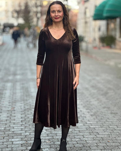 Brun velourkjole med V-hals, trekvart ermer, A-linjeform. Kvinne står på brosteinsgate.