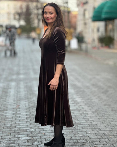Kvinne i brun velourkjole med V-hals og trekvart ermer, står på brosteinsgate med svarte sko.