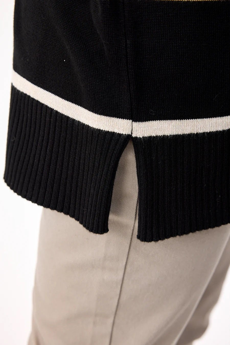 Nærbilde av genserens ribbestrikkede kant med splitt og kontraststripe, over lyse bukser.
