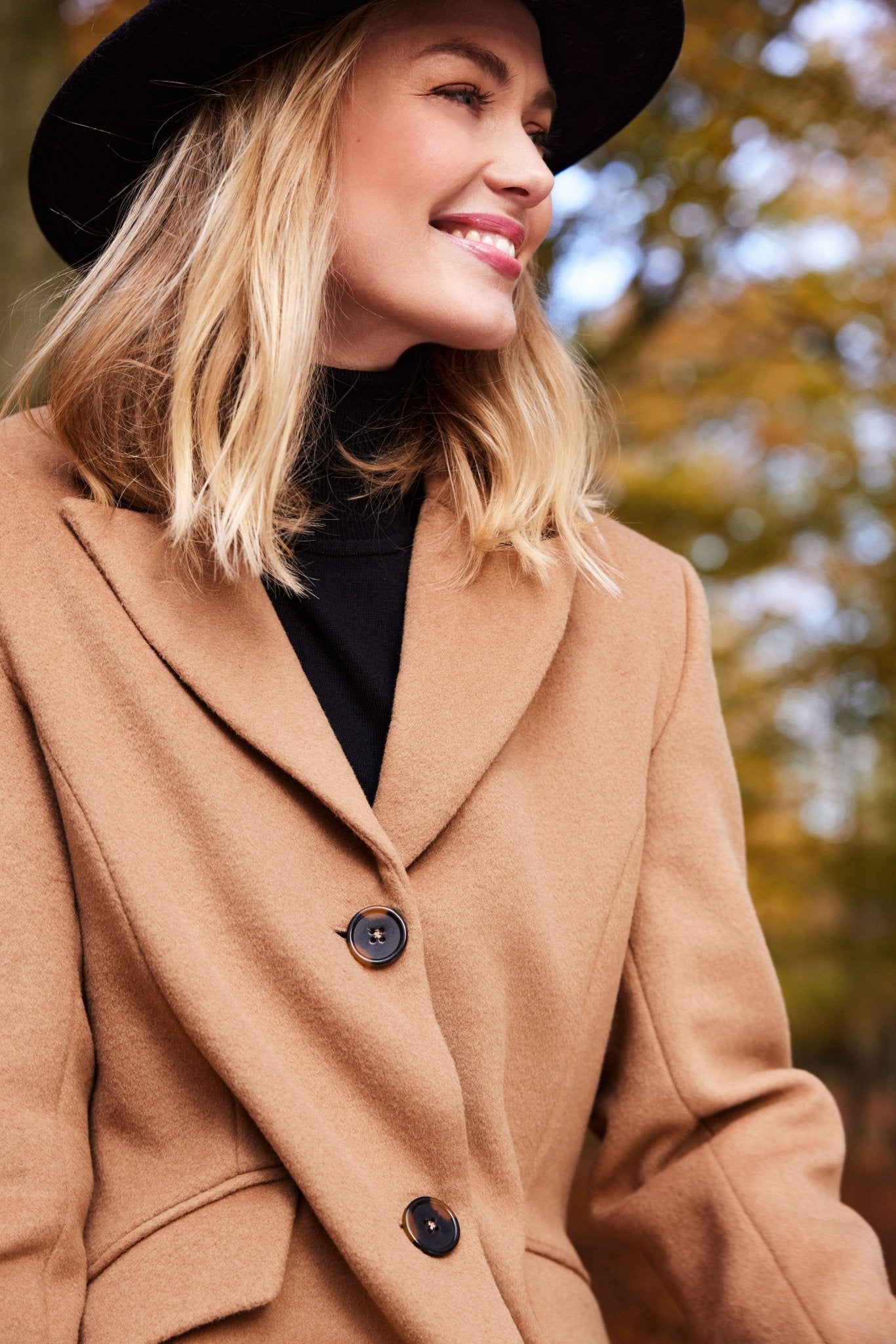 Brun ullkåpe med klassisk krage og to knapper, båret av smilende person i skog.