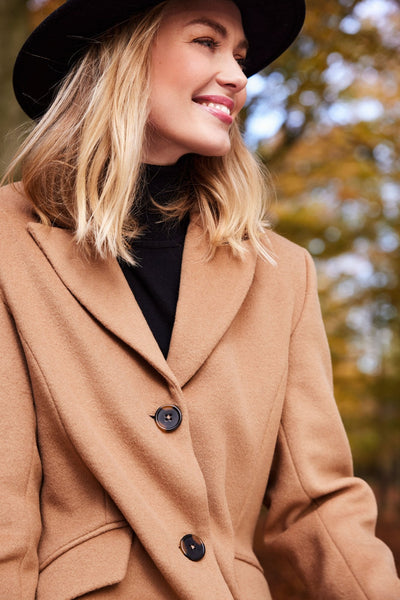 Brun ullkåpe med klassisk krage og to knapper, båret av smilende person i skog.