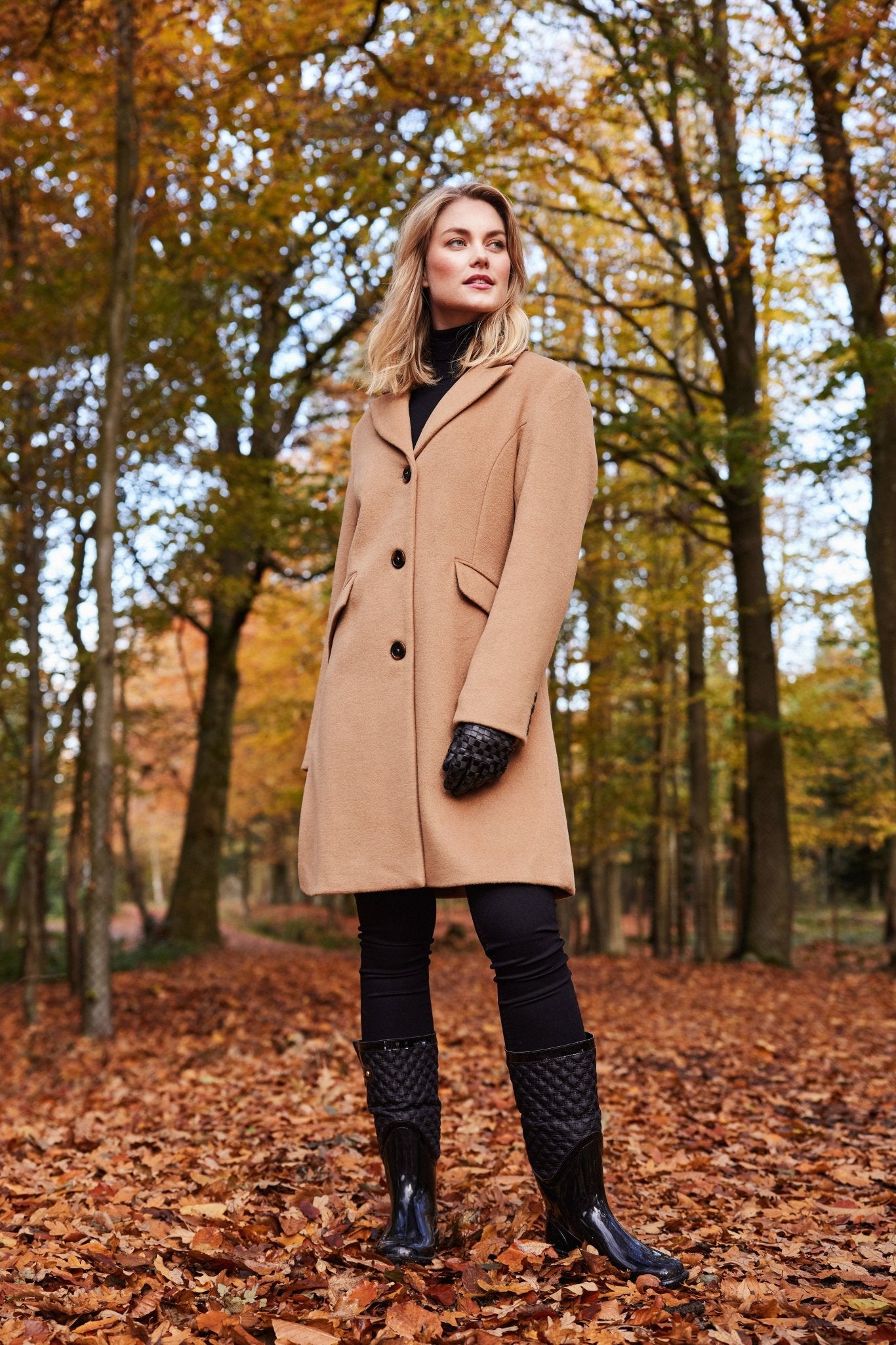 Person i brun ullkåpe med tre knapper og lommer, står i en høstskog.