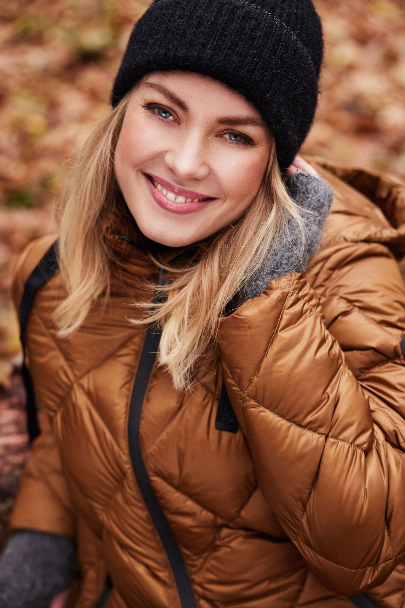 Kvinne i brun, quiltet dunkåpe med glidelås og svart lue, smilende i høstomgivelser.