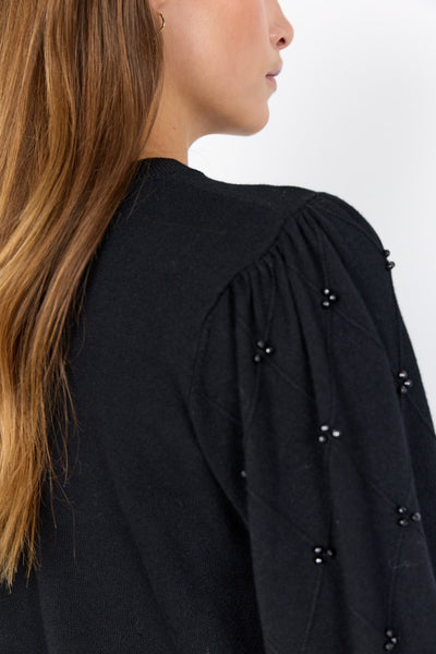 Nærbilde av svart genser med perledetaljer på ermet, sett bakfra. Langt, brunt hår vises.