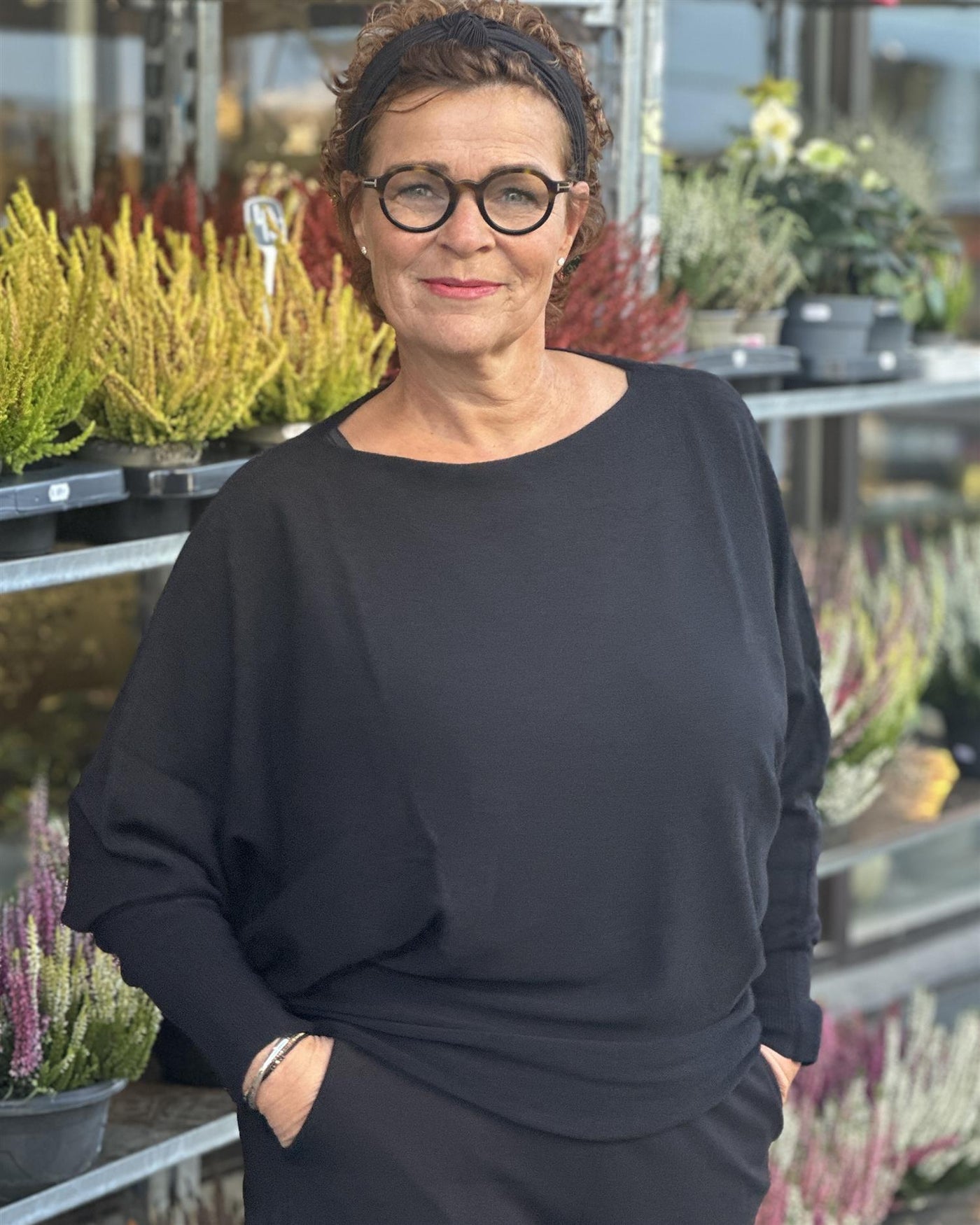 Kvinne iført svart genser med løs passform og bred hals, omgitt av planter utendørs.