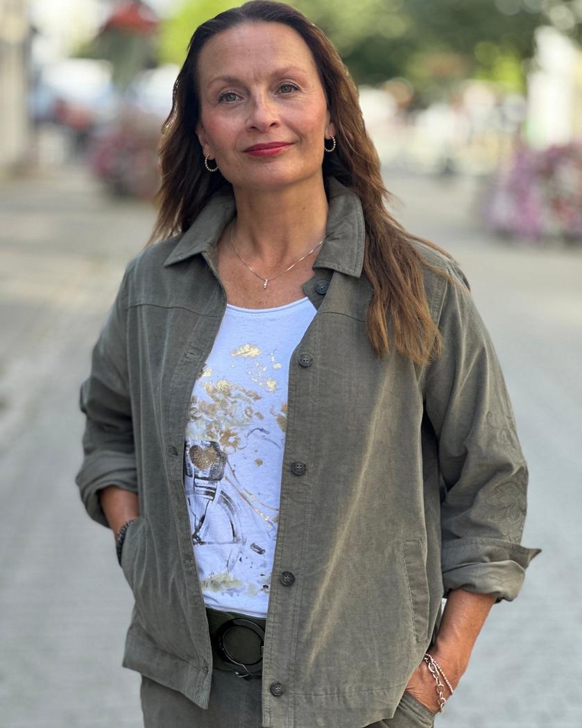 Grågrønn skjorte med knapper og broderte ermer, bæres av en kvinne på en gate.