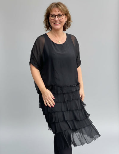 Kvinne i svart, lagdelt kjole med frynser, løs passform og korte ermer, står utendørs.