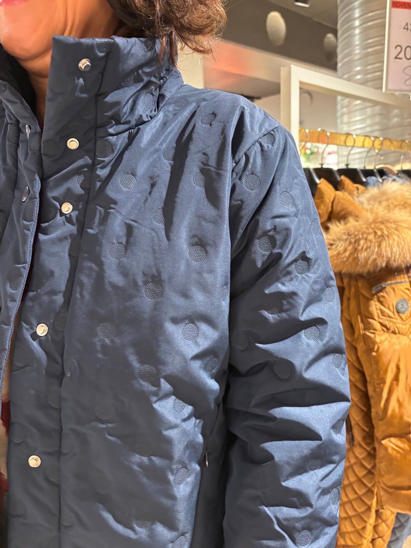 Mørkeblå jakke med sirkulært mønster og sølvknapper, bakgrunn med andre jakker i butikk.
