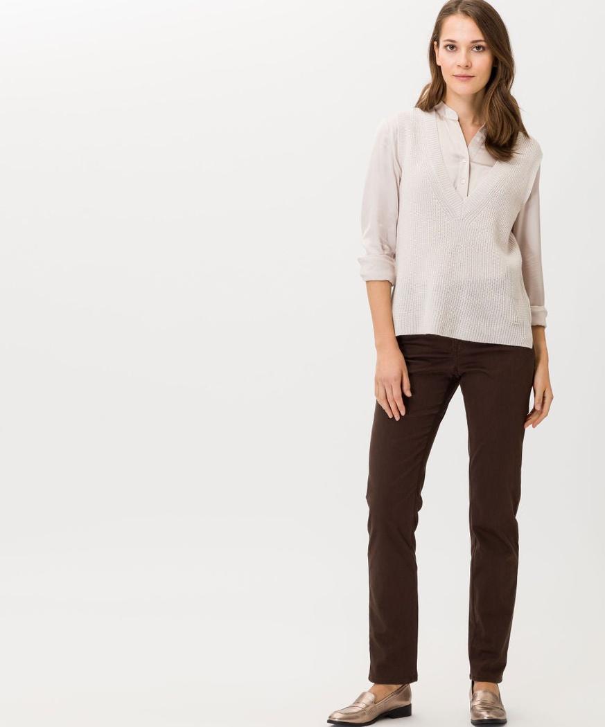 Person i lys bluse, beige strikkevest, mørkebrune bukser og gullsko, står mot lys bakgrunn.