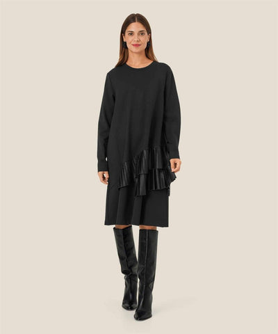 Kvinne i sort kjole med lange ermer og asymmetrisk rysjedetalj. Kjolen rekker til knærne.