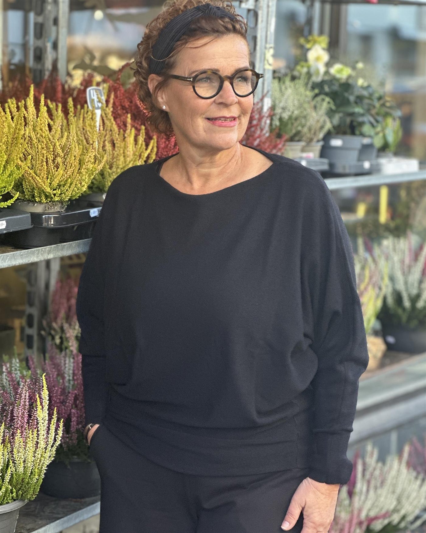 Kvinne i svart genser med bred hals og lange ermer, omringet av planter i et uterom.