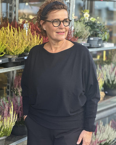 Kvinne i svart genser med bred hals og lange ermer, omringet av planter i et uterom.