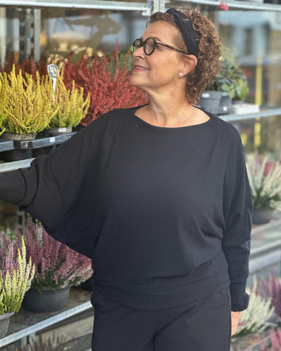 Kvinne i løs svart genser med bred hals, lange ermer, står blant fargerike planter utendørs.