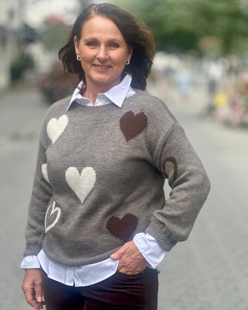 Kvinne med grå genser, hvite og brune hjerter, lange ermer, står utendørs.