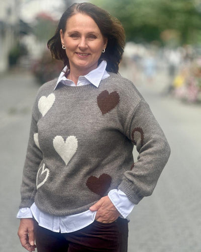 Kvinne med grå genser, hvite og brune hjerter, lange ermer, står utendørs.