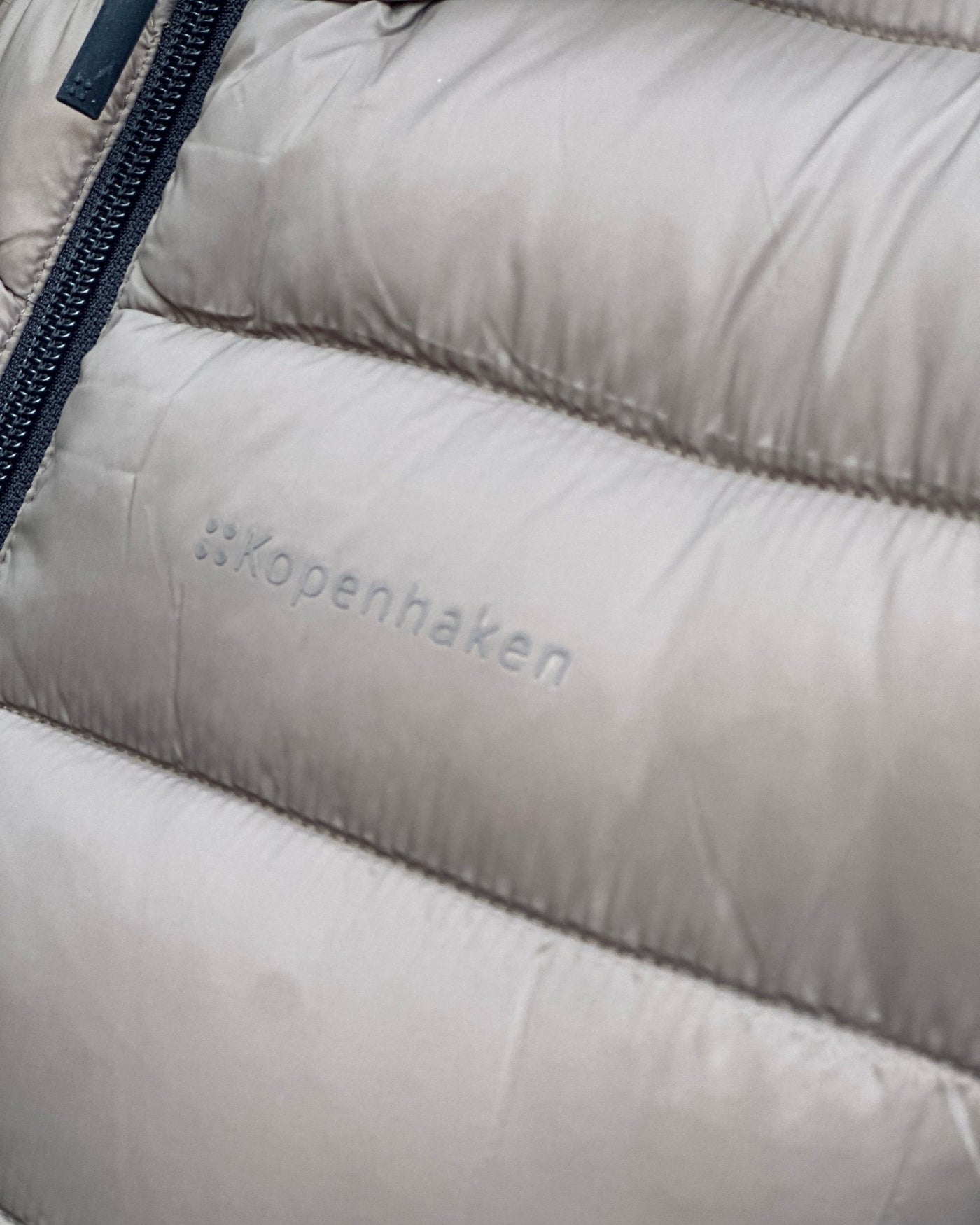 Nærbilde av quiltet jakke med horisontale sømmer, synlig glidelås og "Kopenhaken" logo.