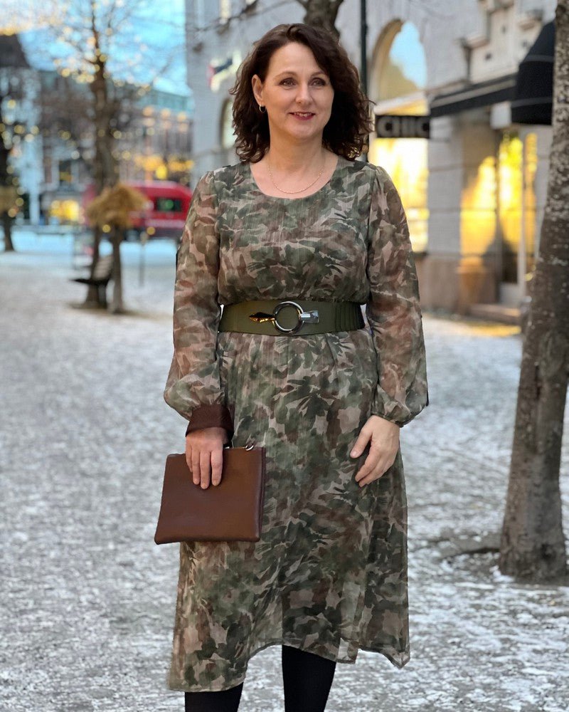 Kvinne i kamuflasjekjole med belte, holder brun clutch på snødekt fortau.