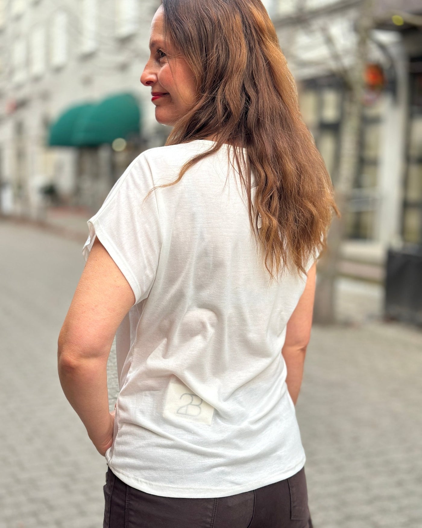 Kvinne i hvit T-skjorte med logo nederst bak, langt brunt hår, står utendørs.