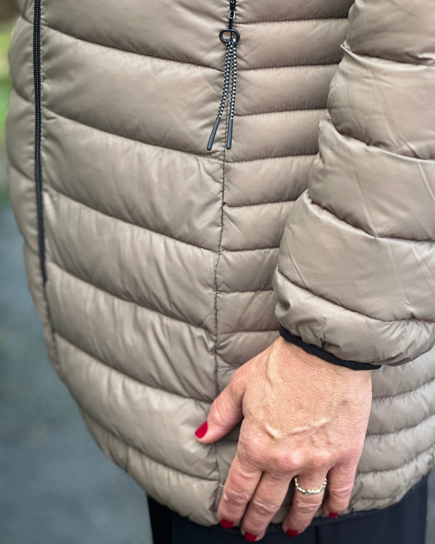 Beige quiltet jakke med glidelås, horisontale sømmer og en hånd med rødlakkerte negler.