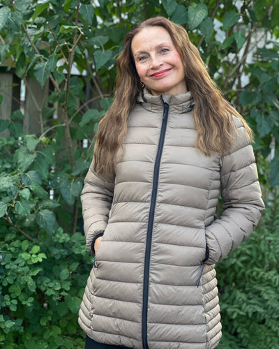 Beige quiltet jakke med hette og glidelås, bæres av kvinne foran grønt løvverk.