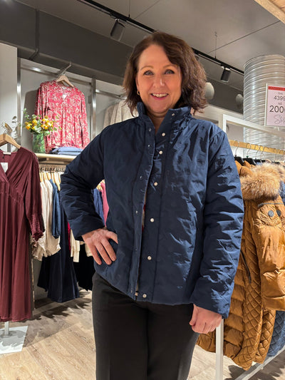 Kvinne i marineblå jakke i butikk, omgitt av fargerike kjoler og jakker på stativ.