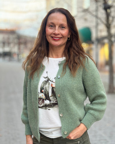 Kvinne med grønn cardigan over en T-skjorte med fargerikt scootertrykk og blomster.