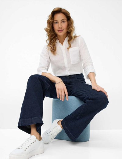 Kvinne i hvit skjorte og mørke jeans sitter på blå krakk, iført hvite sneakers.