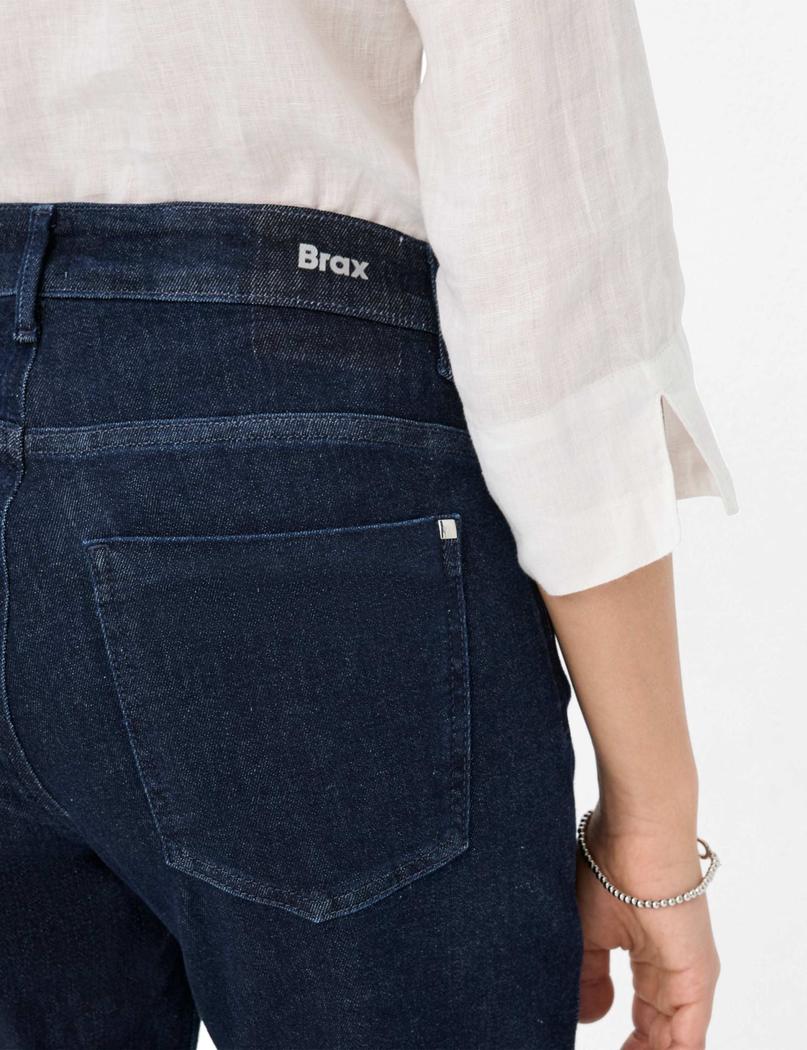 Nærbilde av mørkeblå jeans med "Brax"-logo, hvit skjorte og sølvarmbånd på håndleddet.