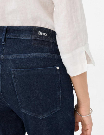 Nærbilde av mørkeblå jeans med "Brax"-logo, hvit skjorte og sølvarmbånd på håndleddet.