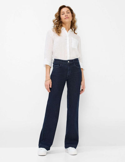 Person med hvit skjorte og mørkeblå vide jeans, står selvsikkert mot hvit bakgrunn.
