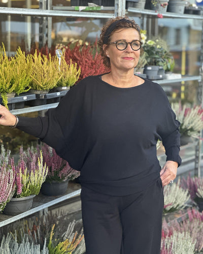 Kvinne i svart genser med løs passform og bred hals, står foran planter i et uterom.