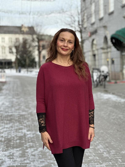 Kvinne i rød, løs genser med splitt og blondedetaljer, trekvart ermer, utendørs bakgrunn.