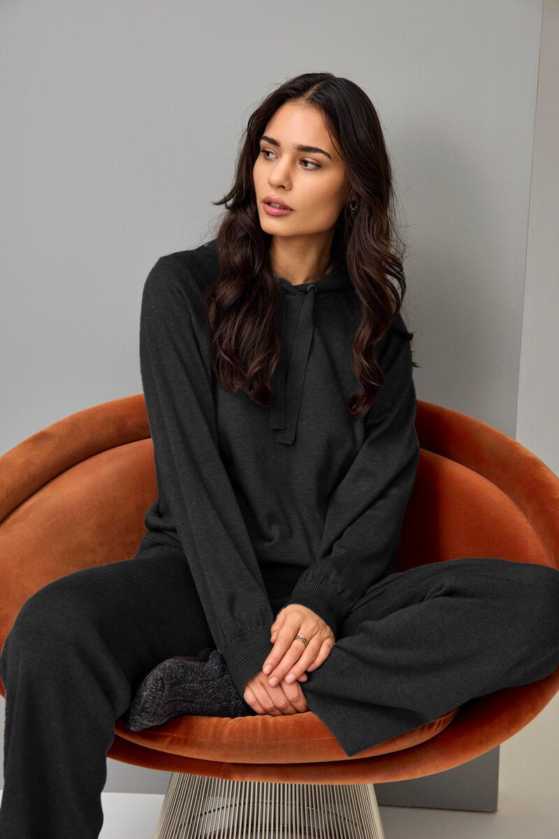 Mørk genser med hette og snøring, lange ermer, avslappet passform, person sitter i stol.