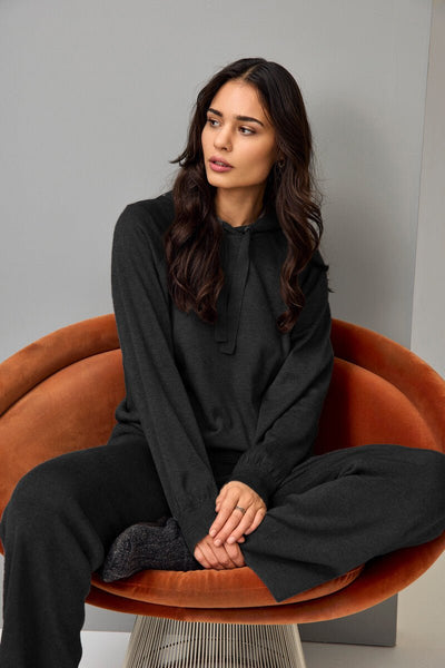 Mørk genser med hette og snøring, lange ermer, avslappet passform, person sitter i stol.