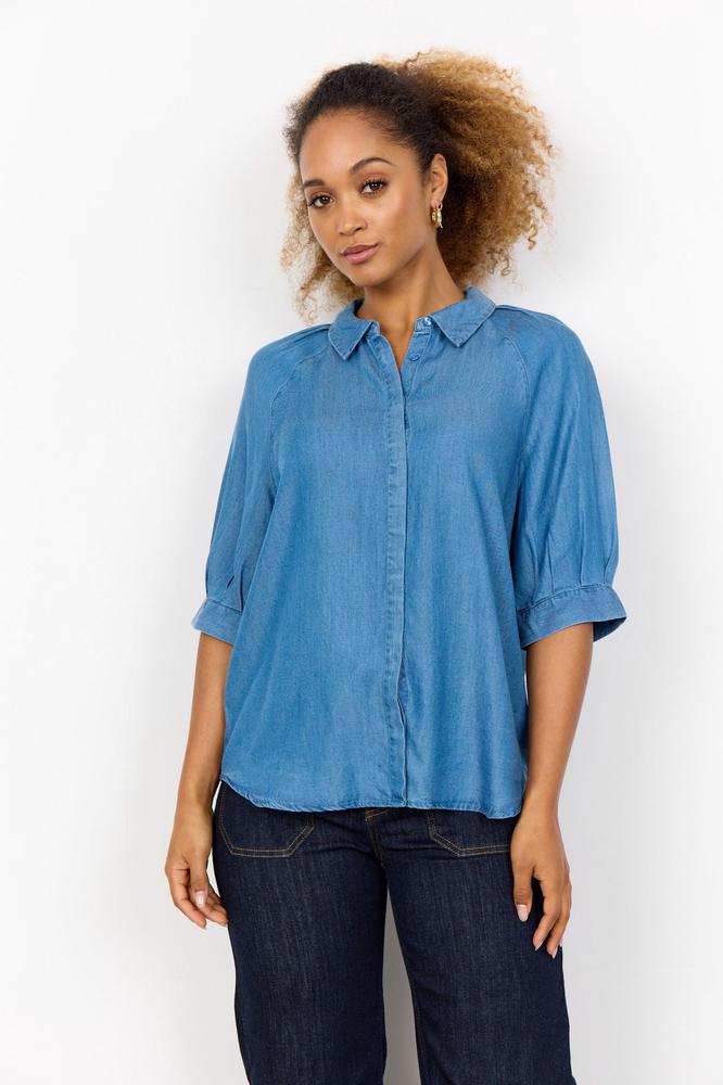 En kvinne iført en blå, avslappet skjorte med oppbrettede ermer og mørke jeans. Hun har krøllete hår.