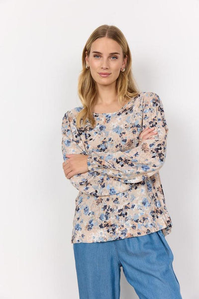 Person med langt, blondt hår iført en bluse med blomstermønster i blå og beige toner, samt blå bukser.
