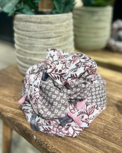 Skjerf med blomstermønster i rosa, grått, hvitt og dusker, på trebord ved siden av en dekorativ potte.