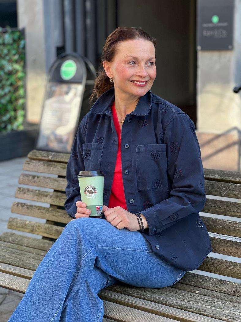 Person på benk med stor, blå brodert skjorte, rød topp, jeans og kaffekopp, smilende.